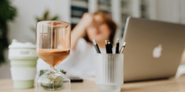 attività invisibili clessidra in prim piano Donna che lavora al pc in secondo piano, con mano sulla fronte. Penso alle attività invisibili che deve fare